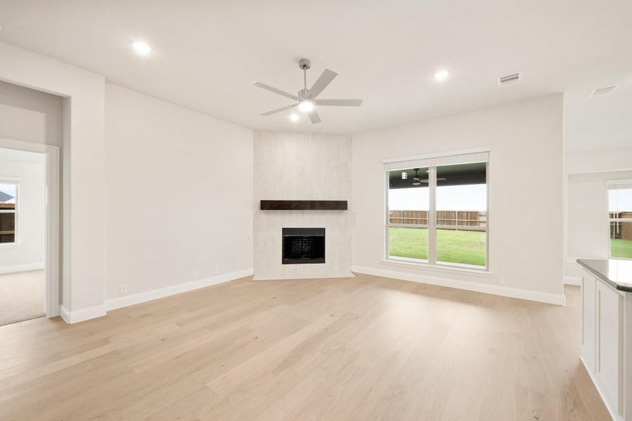 Open living room with light wood floors, corner fireplace, ceiling fan, and large backyard-facing window