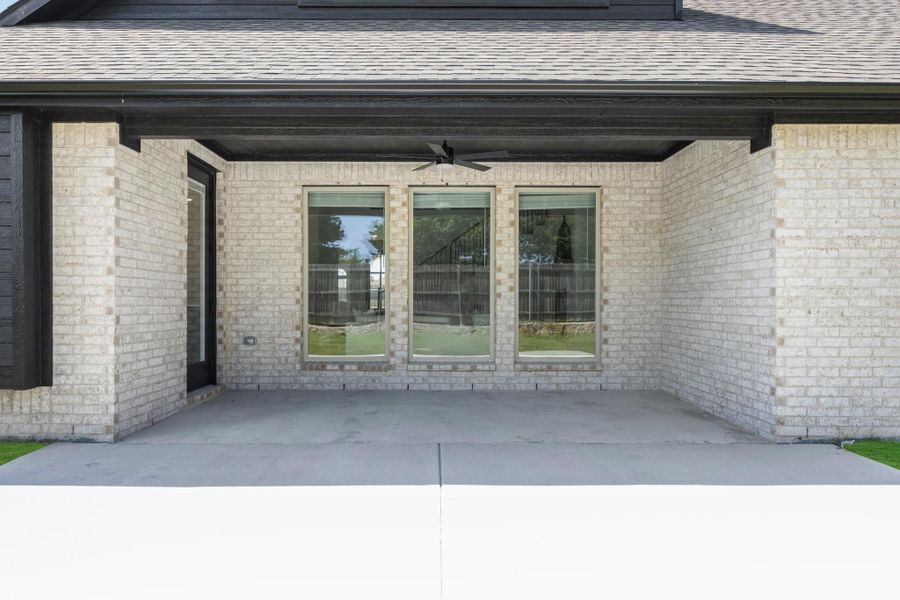Covered back patio with ceiling fan, three large windows, and concrete floor on brick home exterior.