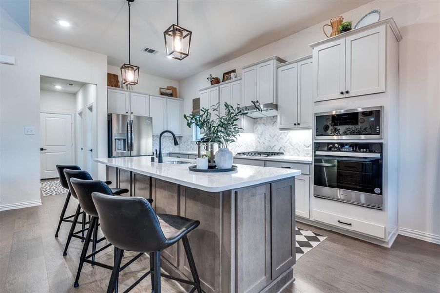Kitchen featuring stainless steel appliances, hanging light fixtures, an island with sink, light stone countertops, and backsplash