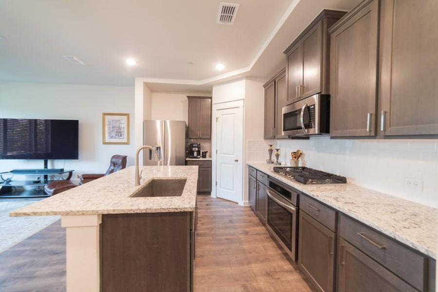 Kitchen featuring appliances with stainless steel finishes, light wood-type flooring, light stone counters, a center island with sink, and backsplash