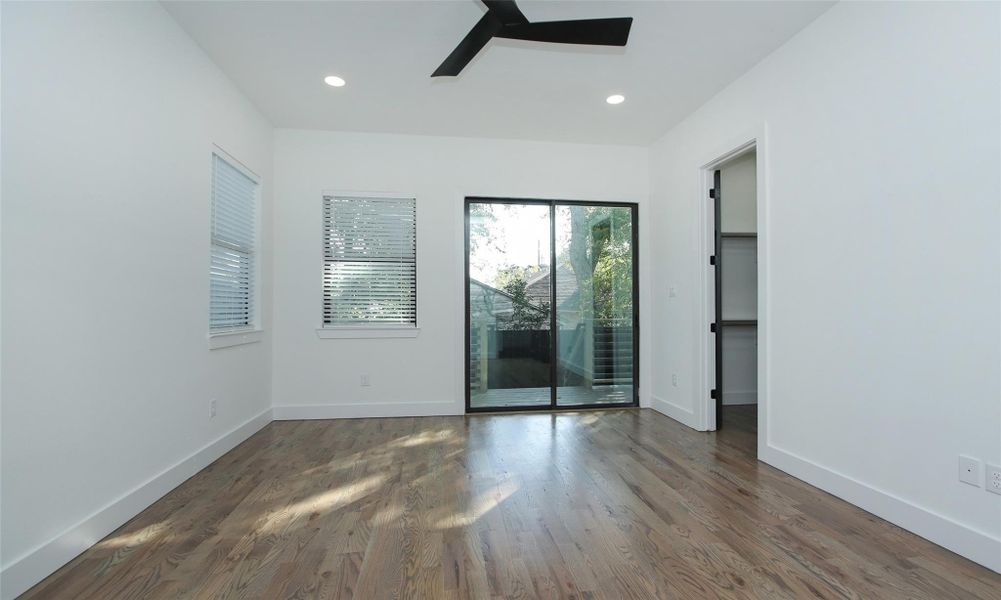 This first floor primary bedroom features hardwood flooring and a modern ceiling fan, with ample natural light from large windows and sliding glass doors leading to a covered back patio. It includes a walk-in closet. This first floor primary bedroom features hardwood flooring and a modern ceiling fan, with ample natural light from large windows and sliding glass doors leading to a covered back patio. It includes a walk-in closet.