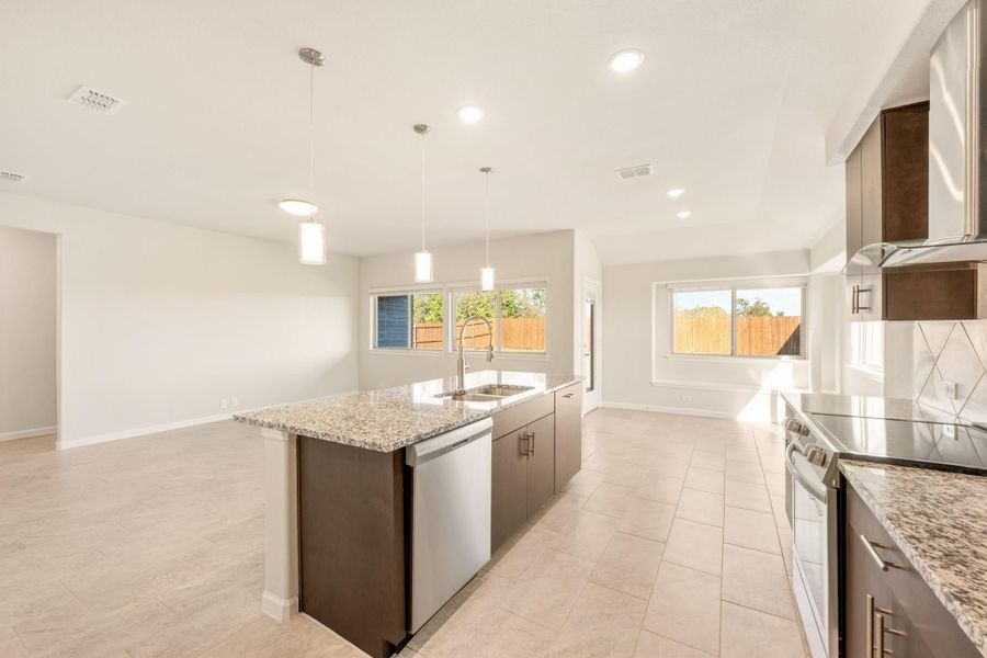Open kitchen with granite island, dark cabinets, pendant lights, and stainless steel appliances opening to living area