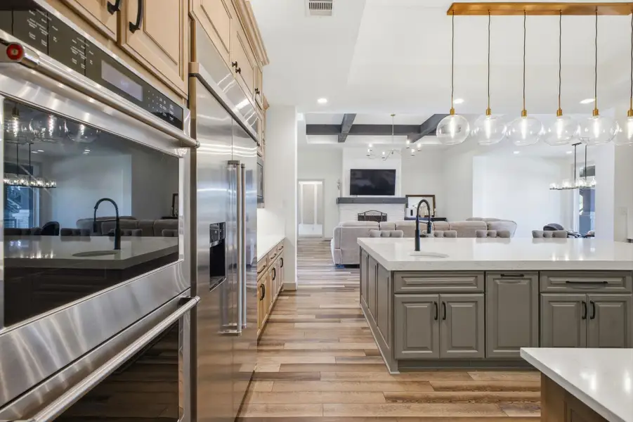 The kitchen features stainless steel appliances, light wood cabinetry, and an expansive island with a contrasting cabinet finish and light countertop, illuminated by unique pendant lighting