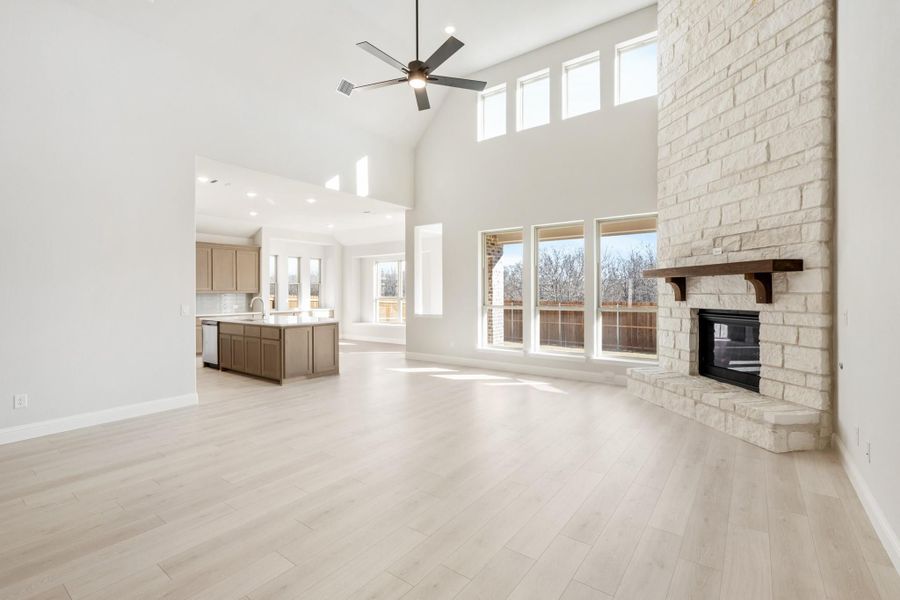 Open living room with vaulted ceiling, stone fireplace, and light wood floors open to kitchen with island
