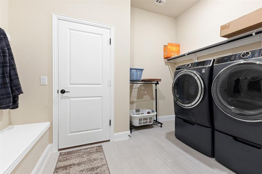 Dedicated laundry area featuring modern front-load appliances on pedestals, overhead shelving, and light-toned tile flooring Dedicated laundry area featuring modern front-load appliances on pedestals, overhead shelving, and light-toned tile flooring