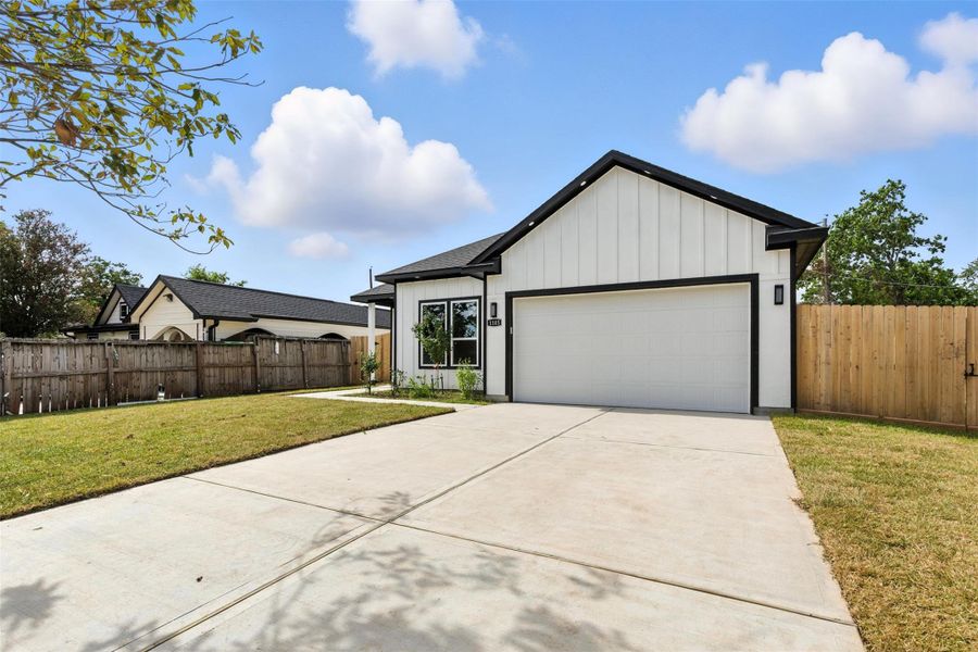 This photo showcases a modern single-story home with a clean white exterior and contrasting black trim. It features a spacious driveway leading to a two-car garage. The property is fenced with a wooden fence, offering a private backyard space. The front yard is well-maintained with a lawn and a few trees.