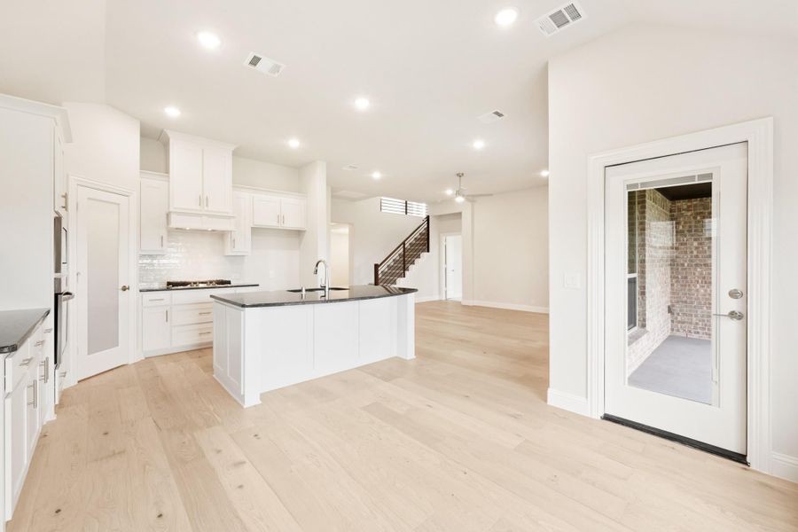 Open-concept kitchen with white cabinets, dark countertop island, and light hardwood floors leading to living area and staircase.
