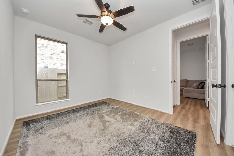 This room features a ceiling fan, wood-like flooring, and a large window for natural light. It has neutral walls, a cozy carpet, and an open doorway leading to another room.