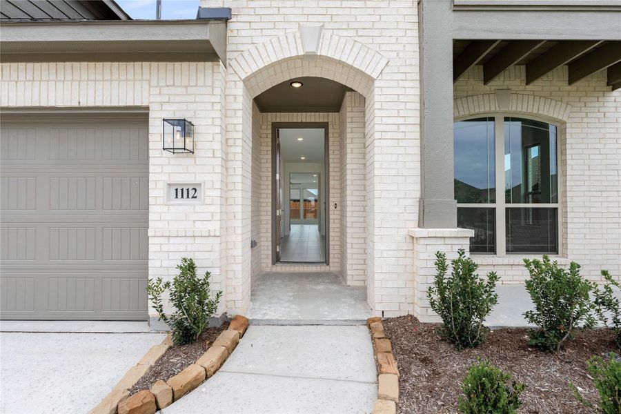 Home entrance with arched brick doorway, address number 1112, single garage door, and landscaped front walkway Home entrance with arched brick doorway, address number 1112, single garage door, and landscaped front walkway