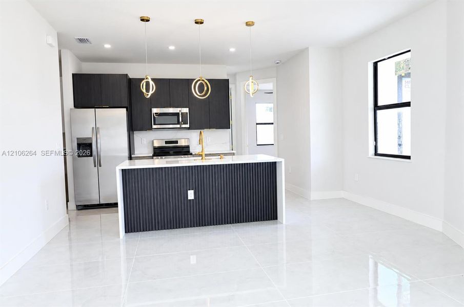 Kitchen featuring a central island with fluted cabinetry and a solid-surface countertop, dark-toned cabinetry, stainless steel appliances, polished tile flooring, and metallic spherical pendant lighting