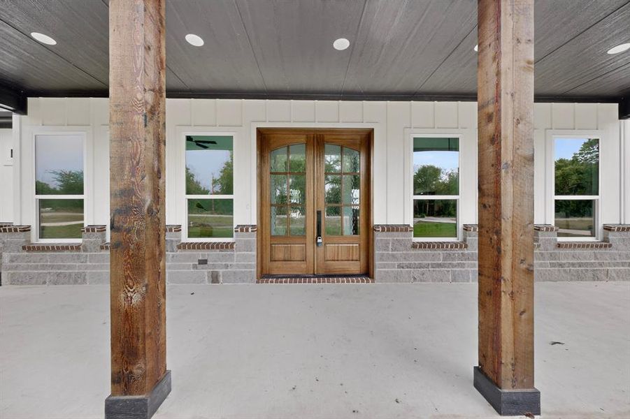 Covered porch with exposed wood-finish columns, a double wood-finish entry door with glass panels, white board-and-batten siding, and a stacked stone foundation with brick accents