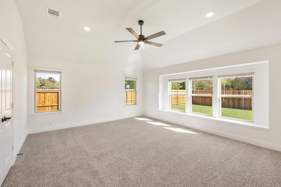 Empty bedroom with vaulted ceiling, ceiling fan, carpet flooring, and large windows overlooking a fenced backyard Empty bedroom with vaulted ceiling, ceiling fan, carpet flooring, and large windows overlooking a fenced backyard