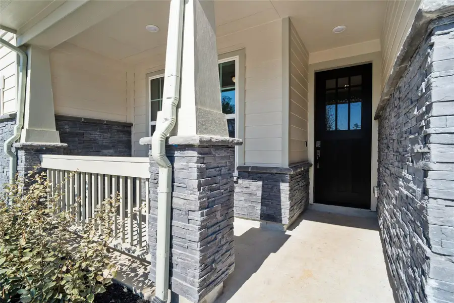 Welcome to a charming, well-maintained front porch, perfect for relaxing and enjoying the outdoors. The porch features sophisticated grey stacked stone accents around the base of the columns and the house's foundation, complemented by neutral beige siding and smooth trim.