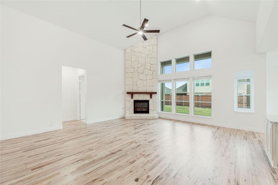 Open living room with tall stone fireplace, hardwood floors, ceiling fan, and large windows
