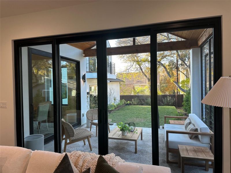 Expansive sliding glass doors open to a covered patio featuring a wood-beam ceiling, concrete flooring, and an outdoor wall sconce