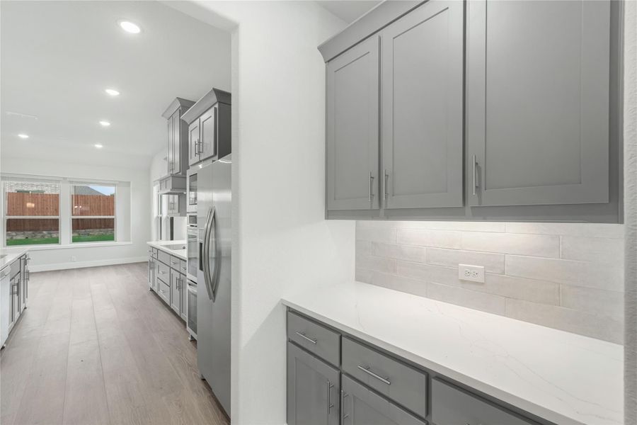 Kitchen with gray shaker cabinets, white quartz countertops, tile backsplash, and stainless steel refrigerator on light wood floors.