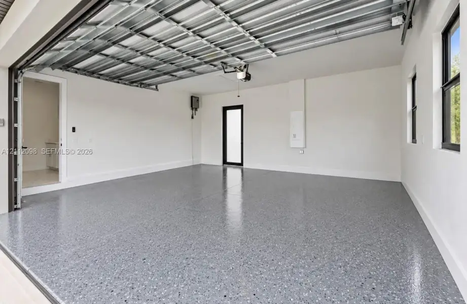 Spacious garage featuring finished epoxy flooring, high-clearance overhead door, smooth-finish walls, and an exterior access door with glass paneling Spacious garage featuring finished epoxy flooring, high-clearance overhead door, smooth-finish walls, and an exterior access door with glass paneling