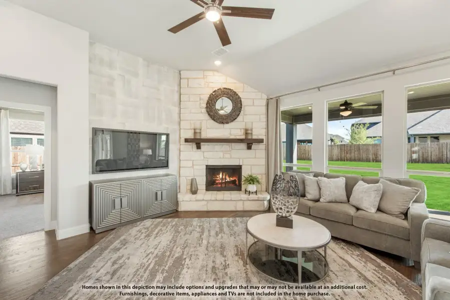 Living room with stone fireplace, vaulted ceiling, ceiling fan, and large windows overlooking backyard