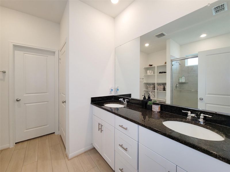 Modern bathroom with dual sinks set in a sleek black countertop, white cabinetry, and a large mirror. The room features light wood flooring, recessed lighting, and a walk-in shower with glass doors. Ample storage space with open shelving enhances functionality.