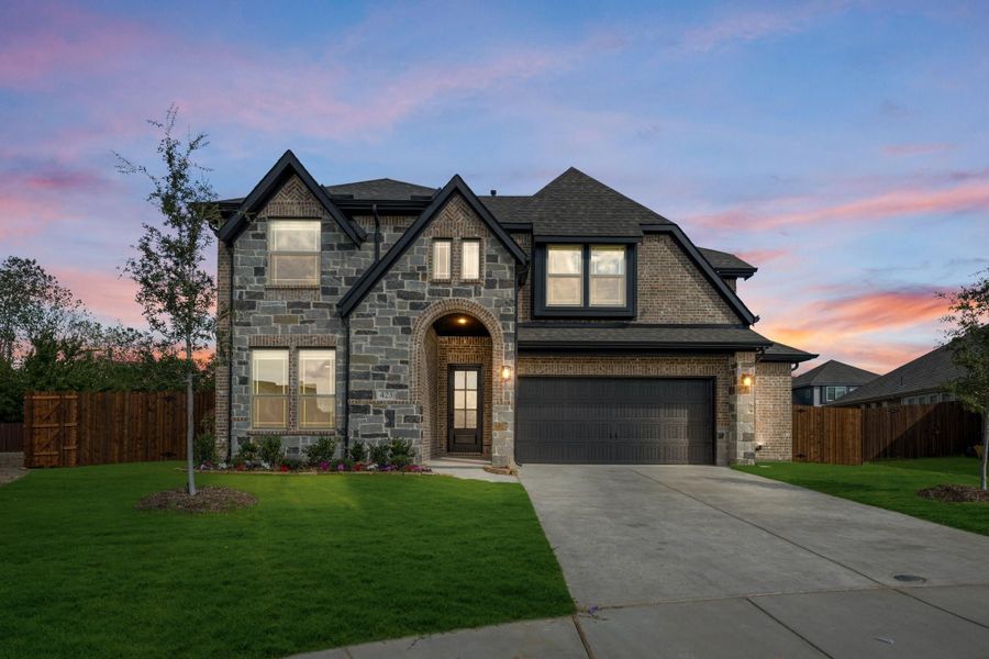 Two-story home with stone and brick exterior, arched entry, two-car garage, and green front lawn at dusk