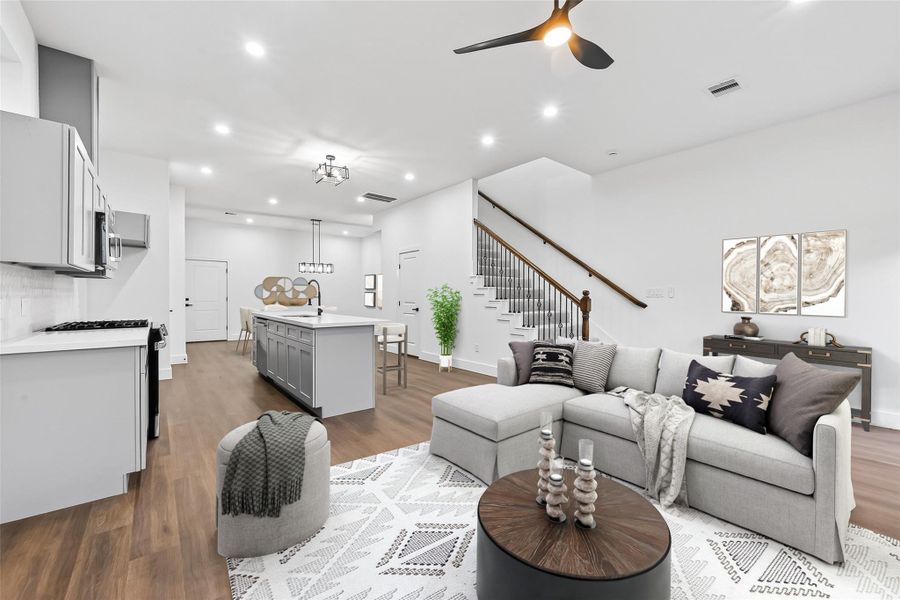 This open-concept living space features a modern kitchen with an island, sleek cabinetry, and stainless steel appliances. The adjacent living area offers a cozy sectional, stylish decor, and a ceiling fan. Natural light brightens the room, highlighting the elegant staircase and hardwood floors.