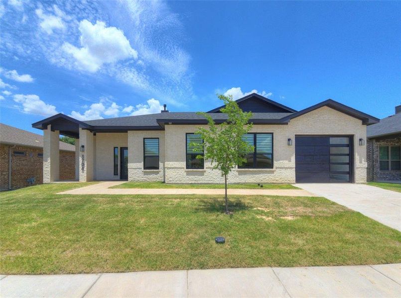 Contemporary single-story residence featuring light-toned brick and stucco facade, a dark-pitched roofline, a covered entry with columns, a modern garage door with frosted glass panels, and a concrete driveway