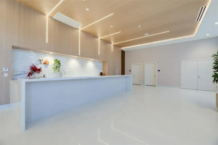 Large kitchen island with a white waterfall countertop, expansive wood-paneled wall with integrated linear lighting, stone-tiled backsplash, polished tile flooring, and recessed ceiling lighting