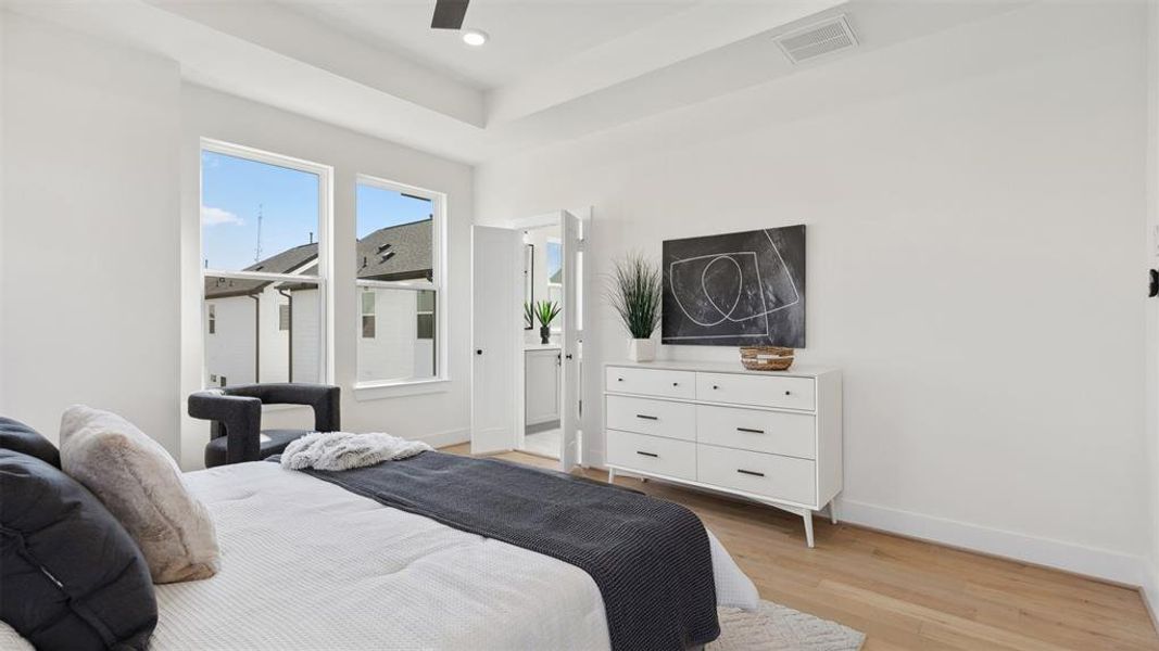 This bright, modern bedroom features large windows, a cozy bed, and sleek furnishings, including a white dresser and contemporary artwork. The space has a clean, minimalist aesthetic with light wood flooring and neutral colors.