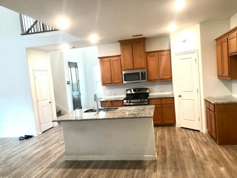 Kitchen with brown cabinets, light stone counters, stainless steel appliances, dark wood-style flooring, and a center island with sink Kitchen with brown cabinets, light stone counters, stainless steel appliances, dark wood-style flooring, and a center island with sink