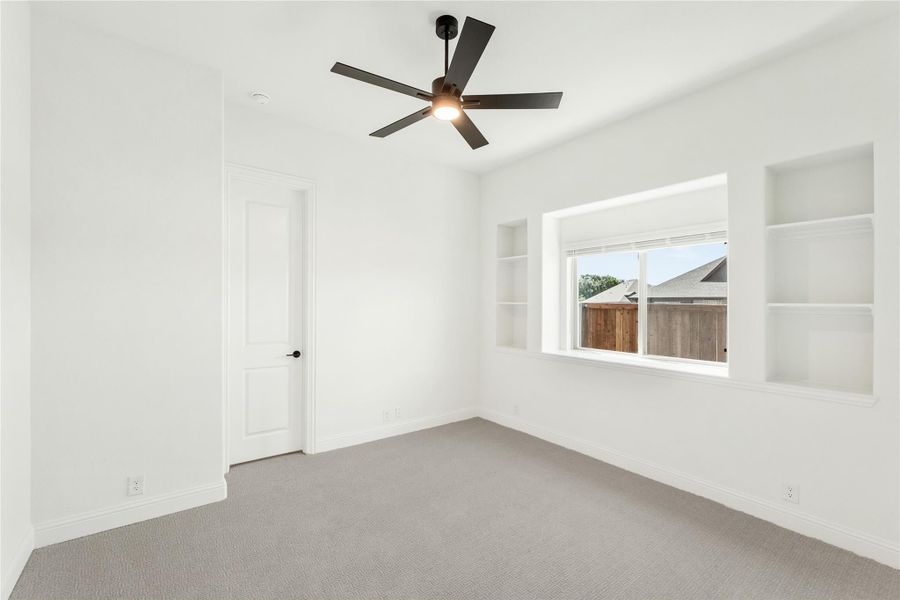 Empty bedroom with gray carpet, ceiling fan, built-in shelves, and a window with backyard view