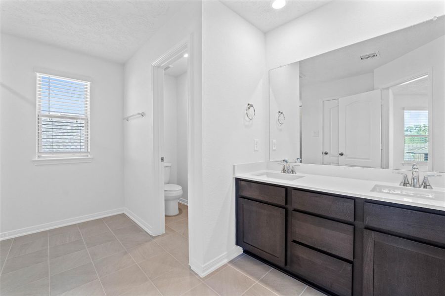 The primary bathroom is move-in ready! It showcases stained cabinetry paired with light countertops, dual vanity sinks, a large mirror, a spacious walk-in closet with built-in shelving, toilet compartment, high ceilings, custom paint, and stylish modern finishes throughout.