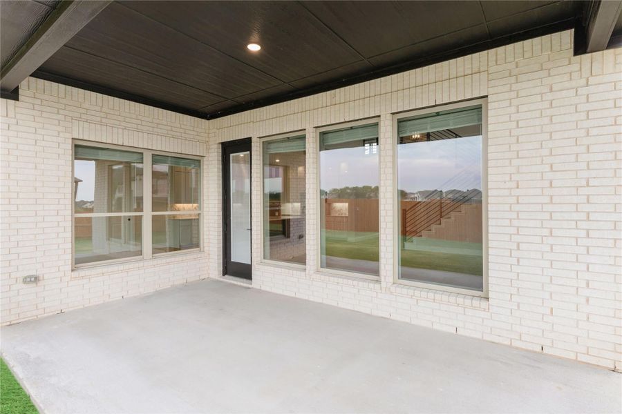 Covered back patio with concrete floor, white brick exterior, large windows, and black door leading inside