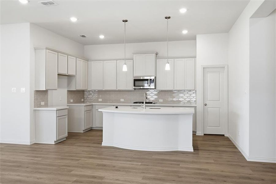 Kitchen featuring a kitchen island with sink, hanging light fixtures, stainless steel microwave, light wood finished floors, and white cabinetry