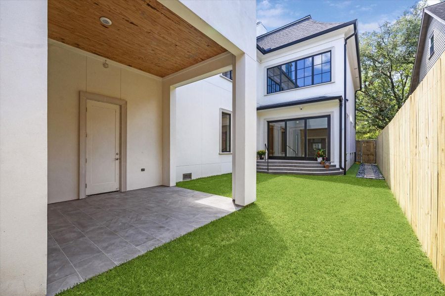 Another view of the new construction, three story home with a clean white exterior, featuring a covered patio, lush green lawn, and a wooden fence for privacy. Large windows and a sliding door enhance natural light.
