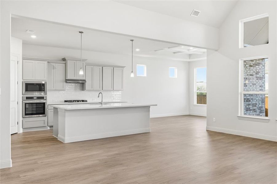 Kitchen featuring appliances with stainless steel finishes, decorative light fixtures, a kitchen island with sink, backsplash, and light wood finished floors Kitchen featuring appliances with stainless steel finishes, decorative light fixtures, a kitchen island with sink, backsplash, and light wood finished floors