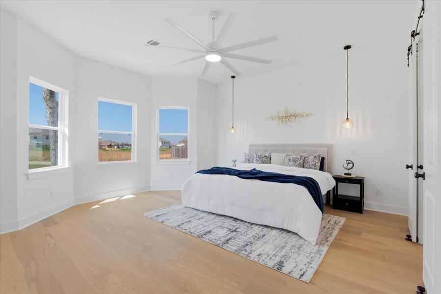 The primary suite features large bay windows that fill the room with natural light, hardwood flooring, a ceiling fan, and elegant pendant lighting on both sides of the bed, each controlled by its own light switch for added convenience.