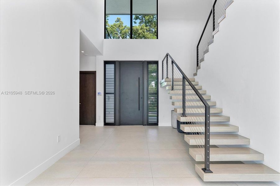 Grand foyer featuring a double-height ceiling, floor-to-ceiling windows, modern floating staircase with cable railing, large-format tile flooring, and a sleek oversized entry door with side lites