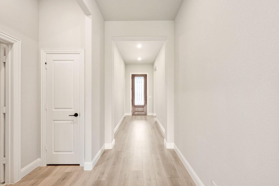 Hallway with white walls, light hardwood floors, and wood front door with glass panel at end Hallway with white walls, light hardwood floors, and wood front door with glass panel at end