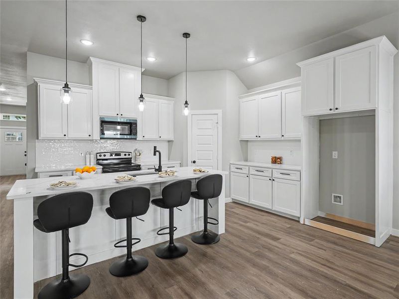Kitchen featuring white shaker cabinetry, an island with countertop seating, wood-finish flooring, a white tile backsplash, and recessed lighting Kitchen featuring white shaker cabinetry, an island with countertop seating, wood-finish flooring, a white tile backsplash, and recessed lighting