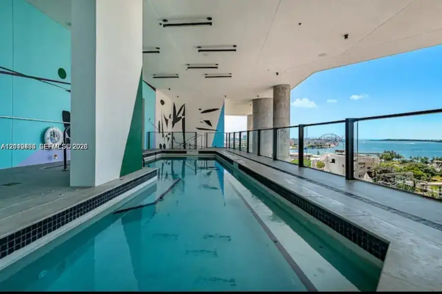 Covered lap pool featuring dark mosaic tile accents, a multi-colored mural wall, recessed ceiling lighting, and a glass panel railing Covered lap pool featuring dark mosaic tile accents, a multi-colored mural wall, recessed ceiling lighting, and a glass panel railing