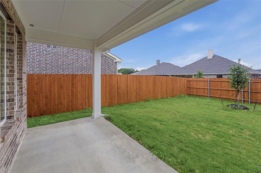 Covered concrete patio with a brick exterior, a wooden privacy fence, a grass lawn, and a young tree