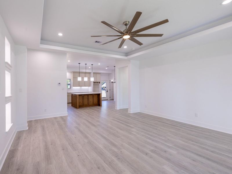 This photo shows a spacious, open-concept living area with light wood flooring, a modern ceiling fan, and recessed lighting. It leads into a kitchen featuring an island with pendant lights and white cabinetry. The space is bright and airy, ideal for entertaining.