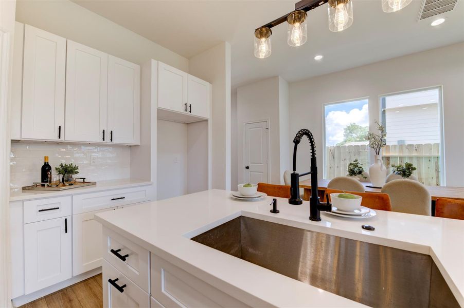 Kitchen island with deep stainless farmhouse sink, matte black coil-spring faucet, quartz countertop, white shaker cabinets, subway tile backsplash, and Edison pendant lighting overhead. Back door with yard view visible.