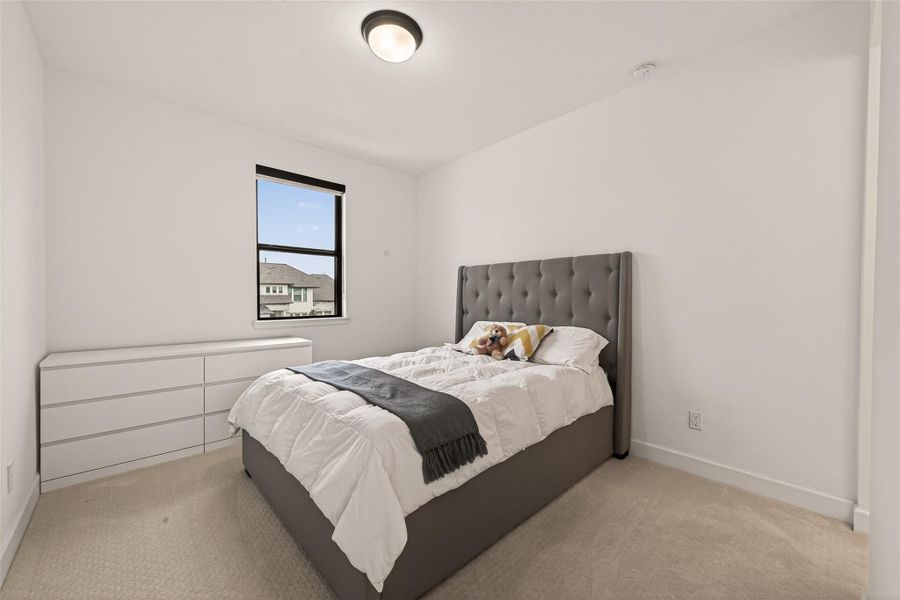 A cozy bedroom featuring a plush, upholstered bed with neutral bedding and accent pillows. The room is bright with natural light from a single window, and there's a sleek, modern dresser. The walls are painted white, enhancing the clean, minimalistic feel.