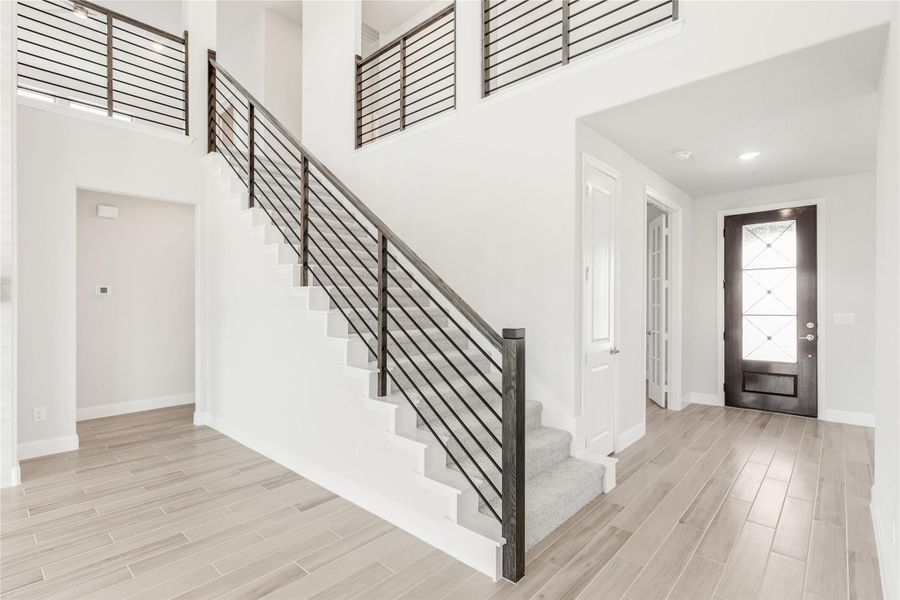 Two-story entryway with staircase, black metal railing, light wood floors, and glass-panel front door Two-story entryway with staircase, black metal railing, light wood floors, and glass-panel front door