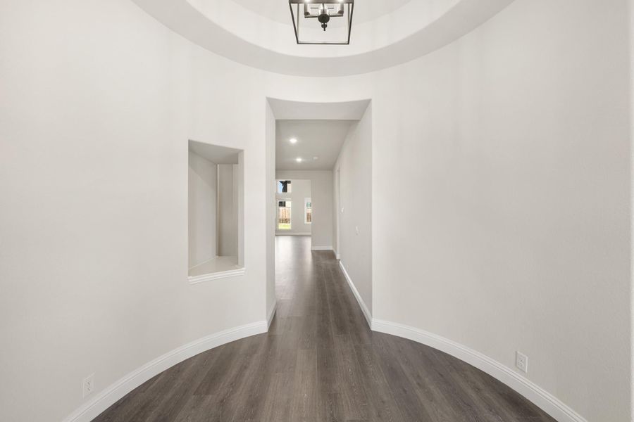 Curved entryway with round tray ceiling, pendant light, dark hardwood floors, and built-in wall niche leading to hallway. Curved entryway with round tray ceiling, pendant light, dark hardwood floors, and built-in wall niche leading to hallway.