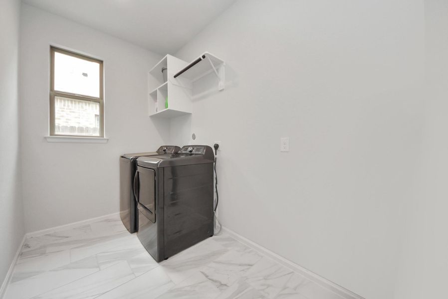 This photo shows a bright, modern laundry room with sleek black washer and dryer units. It features white walls, marble-patterned flooring, and a window for natural light. There's a wall-mounted shelf for storage.