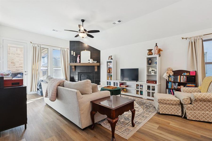 Living room with a tiled fireplace, vaulted ceiling, light wood finished floors, and a ceiling fan Living room with a tiled fireplace, vaulted ceiling, light wood finished floors, and a ceiling fan