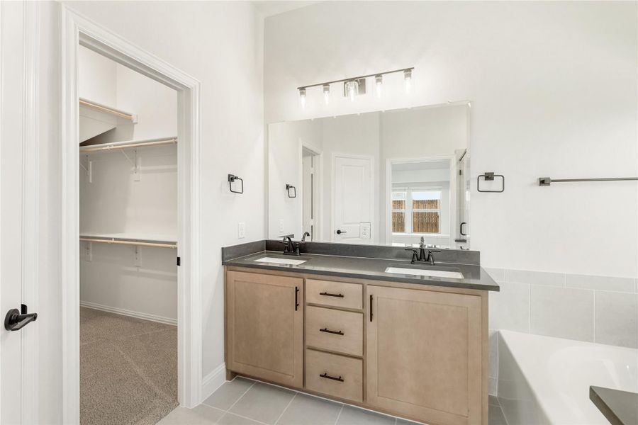 Bathroom with double-sink vanity, wood cabinets, dark countertop, large mirror, and walk-in closet visible through doorway. Bathroom with double-sink vanity, wood cabinets, dark countertop, large mirror, and walk-in closet visible through doorway.