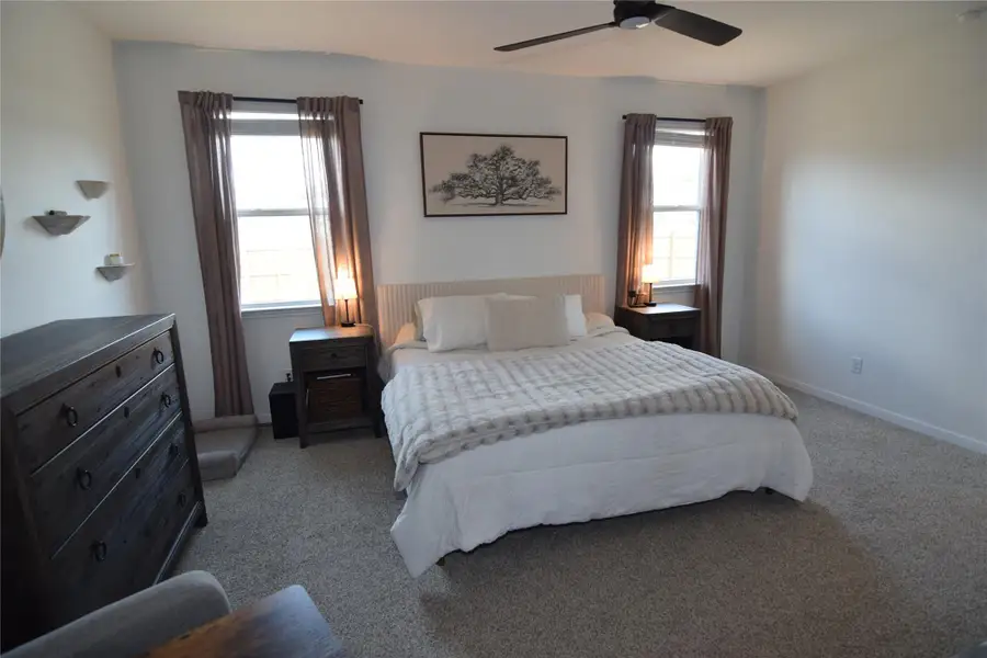 Natural light streams through double windows onto plush carpeted flooring in this comfortable bedroom.